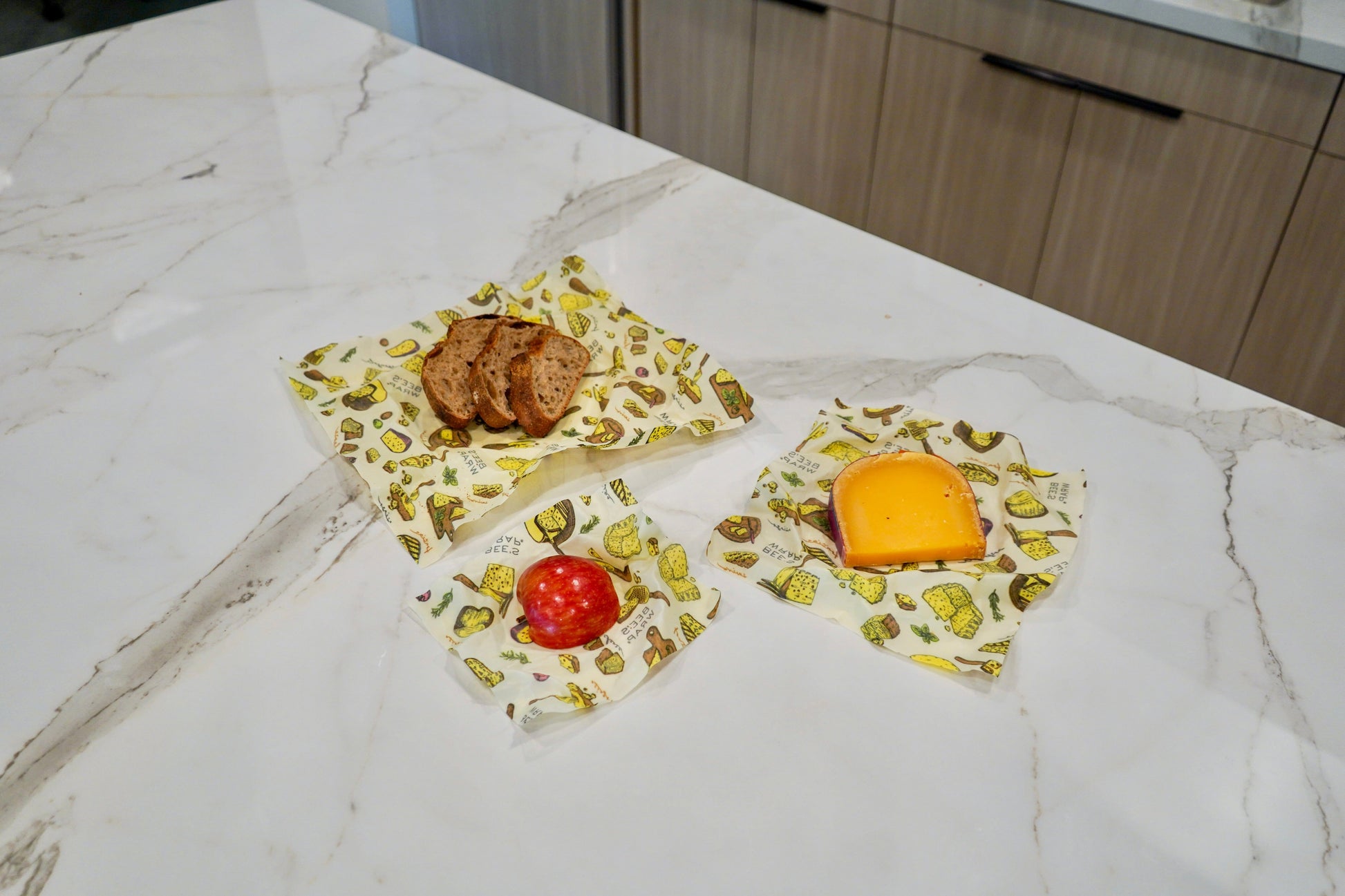 Three reusable Bees Wrap wraps in 3 sizes, small medium large with the Wheels n Wedges print on a marble kitchen countertop, styled with food items (bread, cookies, tomato, cheese) to demonstrate their use.