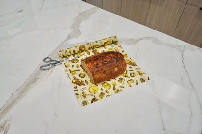 A loaf of bread placed on a reusable Bees Wrap Roll with the Wedges n Wheels print and blue-handled scissors on a white marble kitchen countertop.